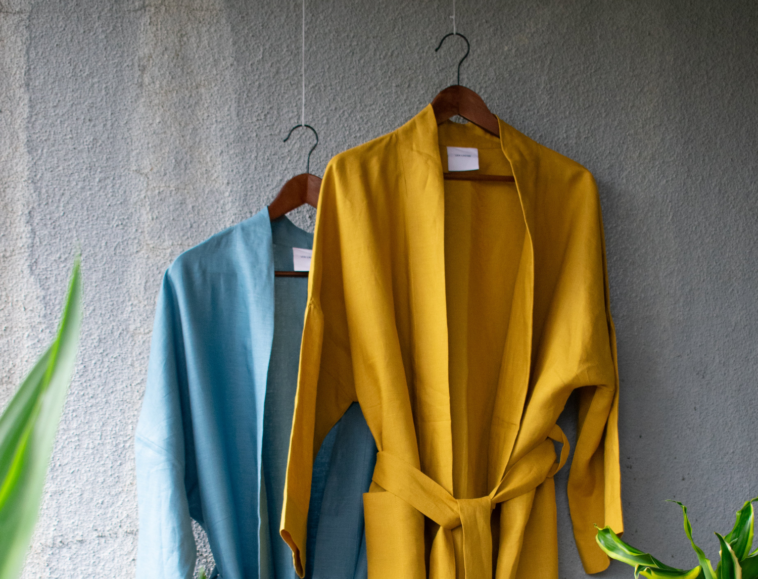 Two summery bathrobe tops hang loosely on hangers blowing in the wind. One in a warm yellow colour, the other in a fresh blue. The tied linen kimono blows loosely. A palm tree adorns the picture.
