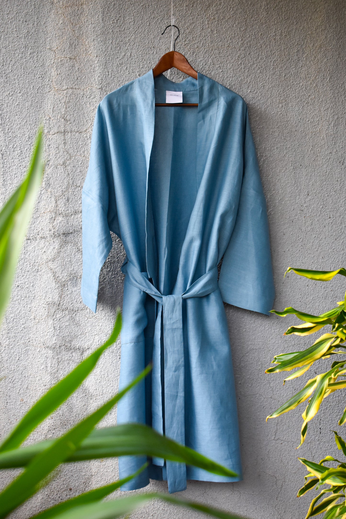 A loose-fitting kimono in a light denim look is ironed on and tied loosely with a wide belt. It reflects a summery look with a green plant at the edge of the picture.
