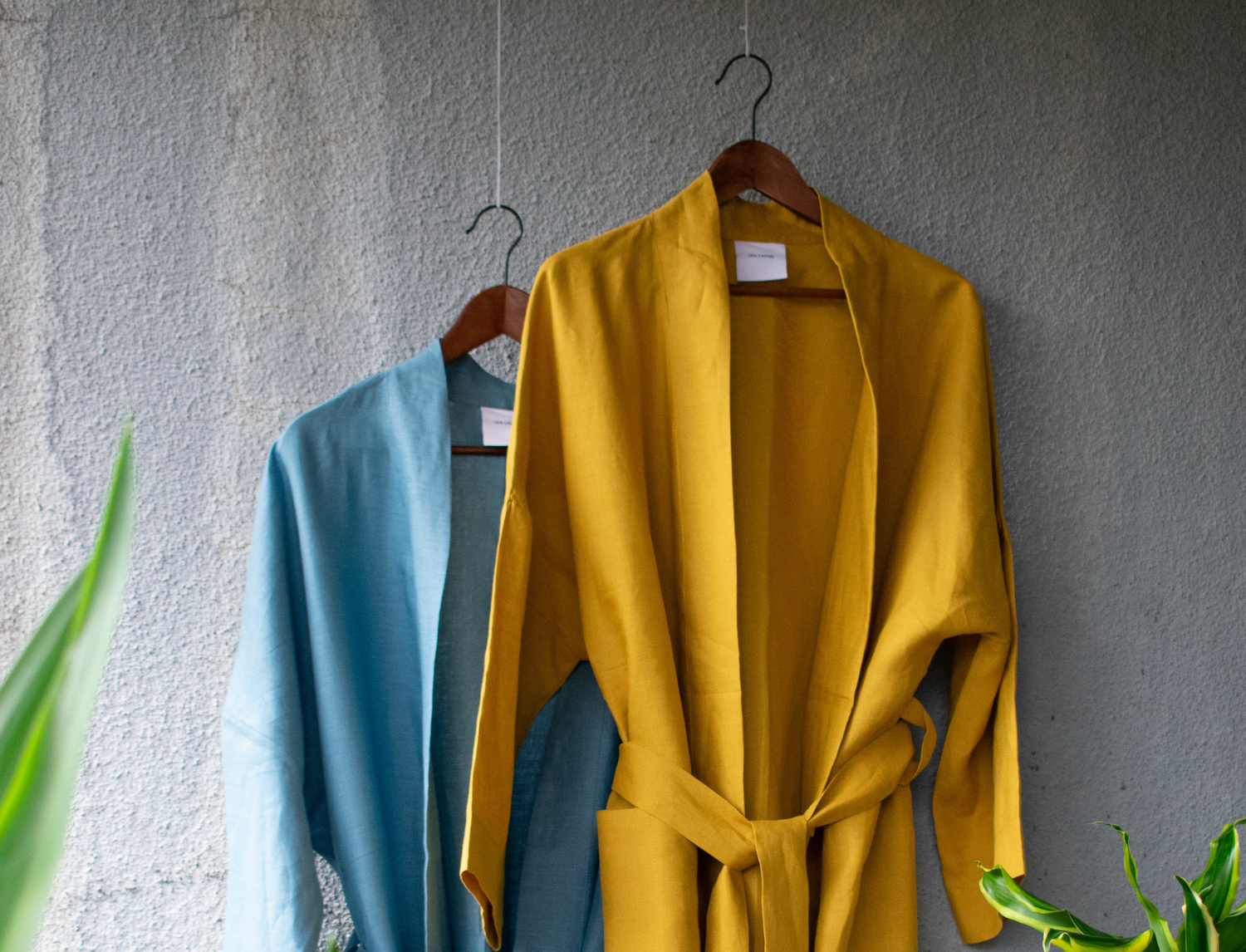 Two summery bathrobe tops hang loosely on hangers blowing in the wind. One in a warm yellow colour, the other in a fresh blue. The tied linen kimono blows loosely. A palm tree adorns the picture.  