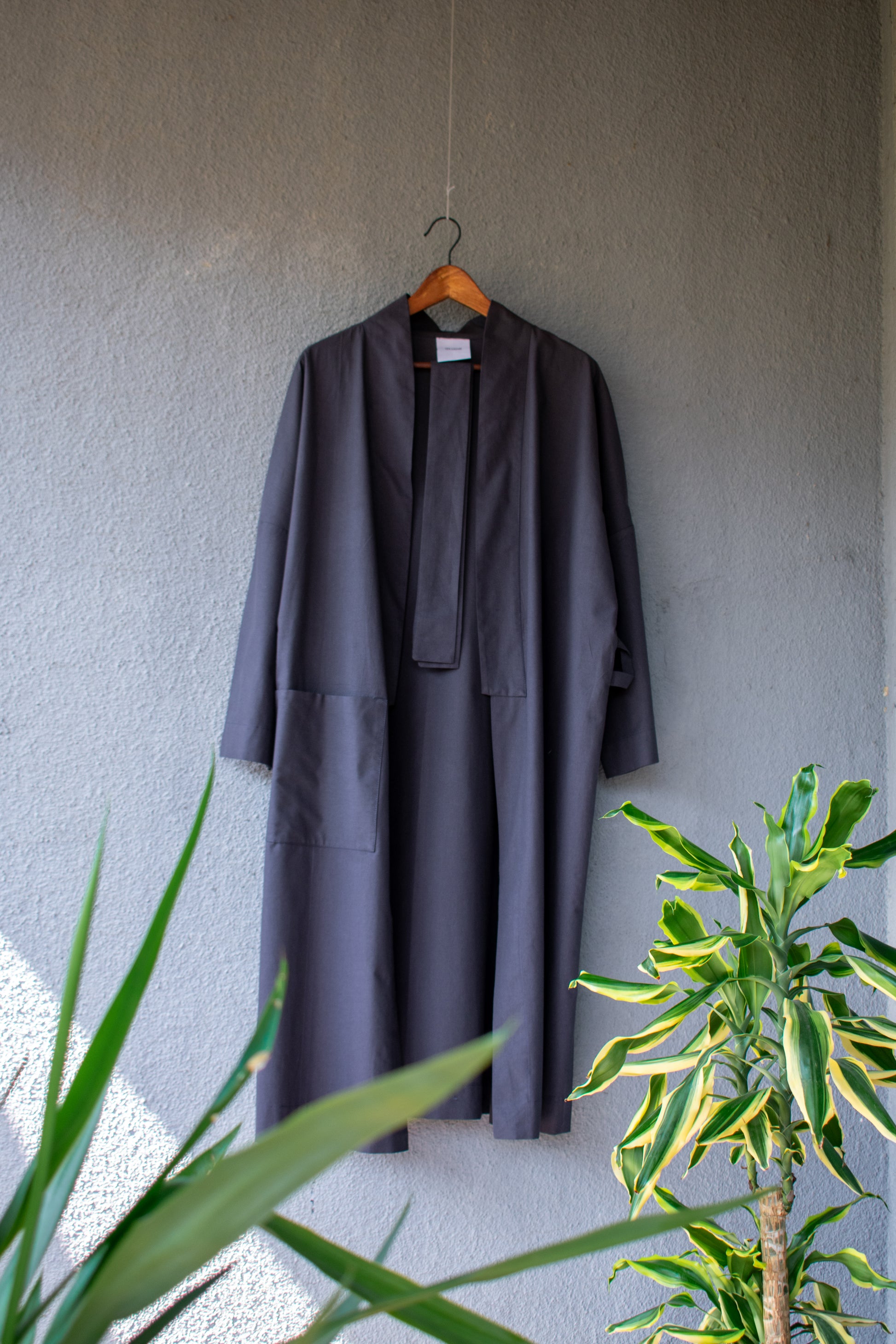 The anthracite-coloured kimono robe hangs open on a hanger. A large side pocket, a long shawl collar and a clean belt emphasise the symmetrical shape of the loose kimono. The firmer cotton fabric gives it a clean drape.