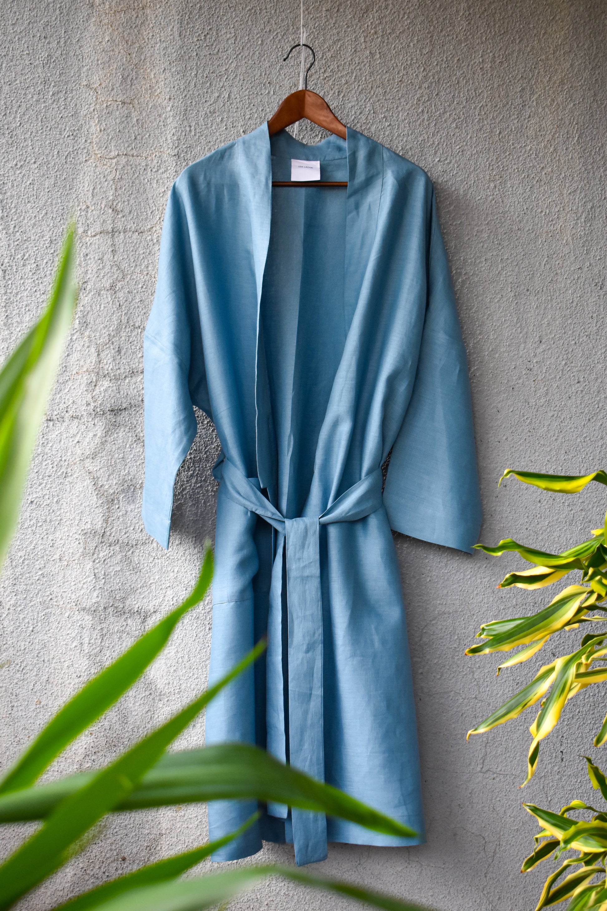 A loose-fitting kimono in a light denim look is ironed on and tied loosely with a wide belt. It reflects a summery look with a green plant at the edge of the picture. 