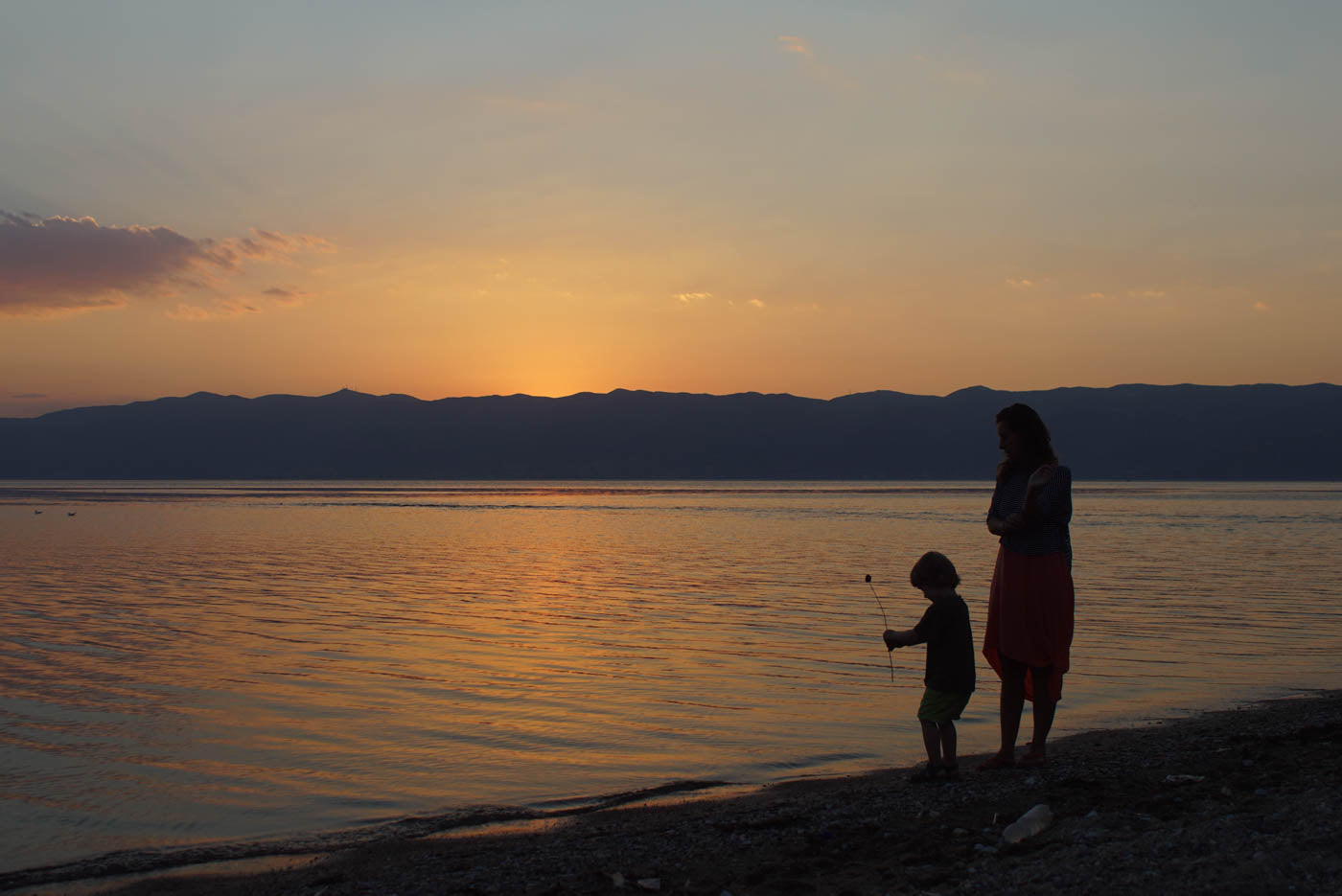 A woman with a playing child stands in the sunset of a large lake. They are enjoying the summery surroundings at sunset and are wearing light clothing.