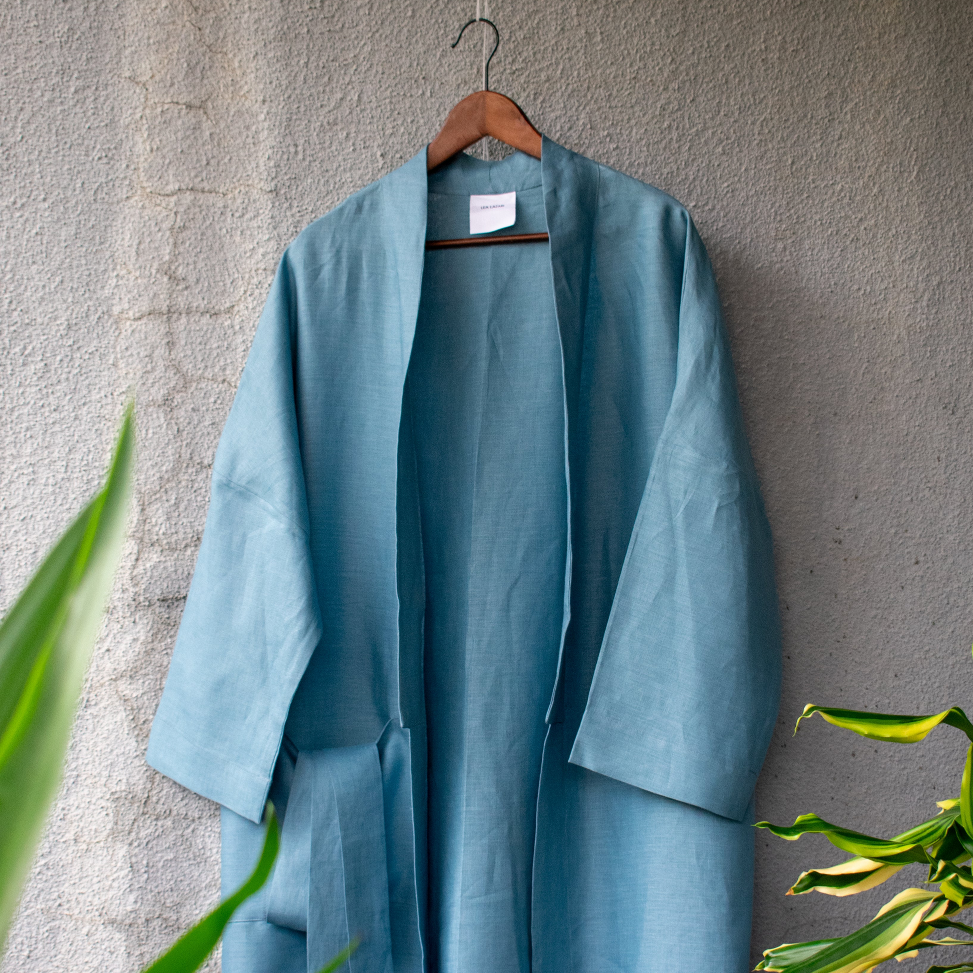 Loose kimono coat blowing in the wind on a light wall with small palm trees in the foreground. It is medium blue, made of fine, smooth summer linen. The coat is open, the belt is in the side pocket.