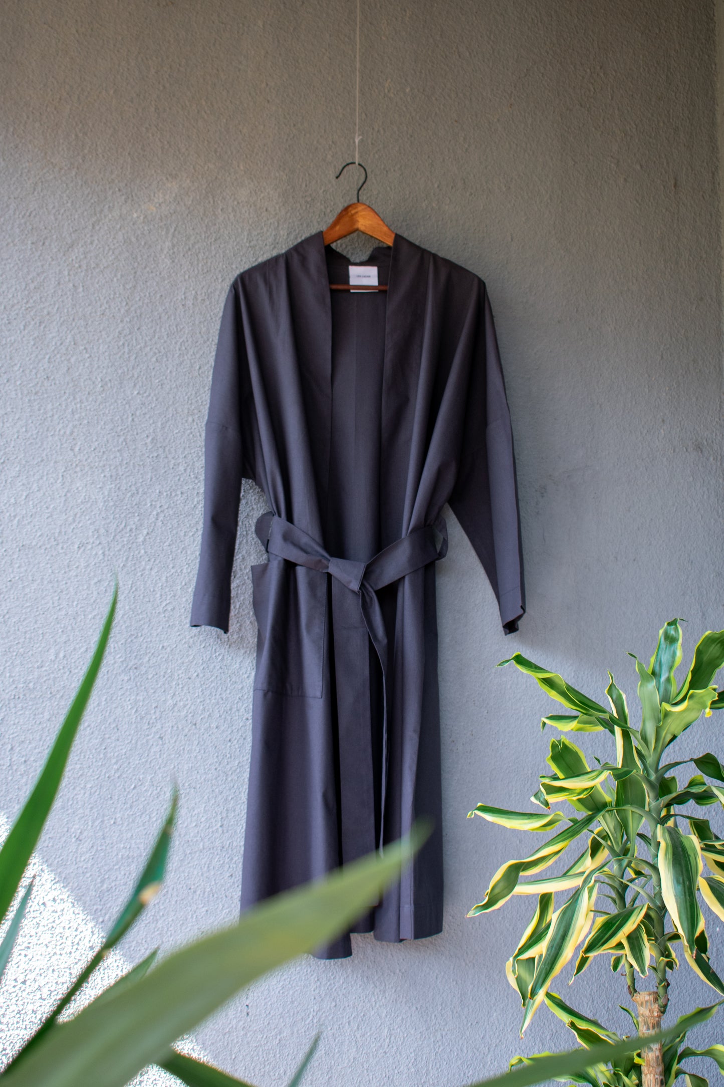 A long cotton kimono hangs loosely on a light-coloured wall. It is wide in shape, tied once and its long sleeves fall loosely. The dark anthracite colour gives the coat a certain elegance.