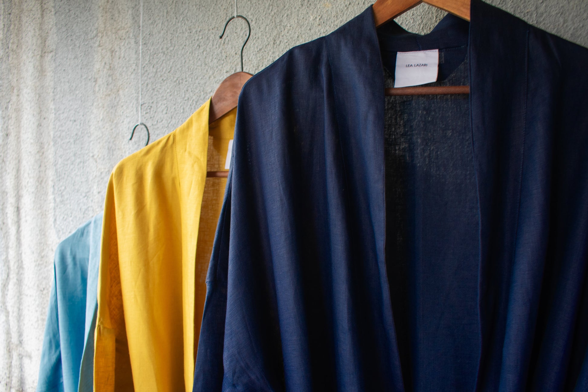 A mustard yellow, a mid-blue and a dark blue summer linen bathrobe hang on three wooden hangers. 