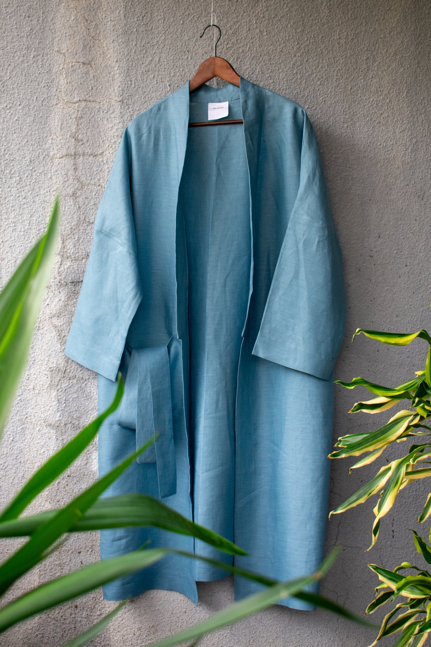 A medium blue loose fit kimono coat swings against a sunny wall with palm trees on the side. The linen looks like very light, soft denim. The coat has a clear, modern shape thanks to its wide collar and a wide slit pocket