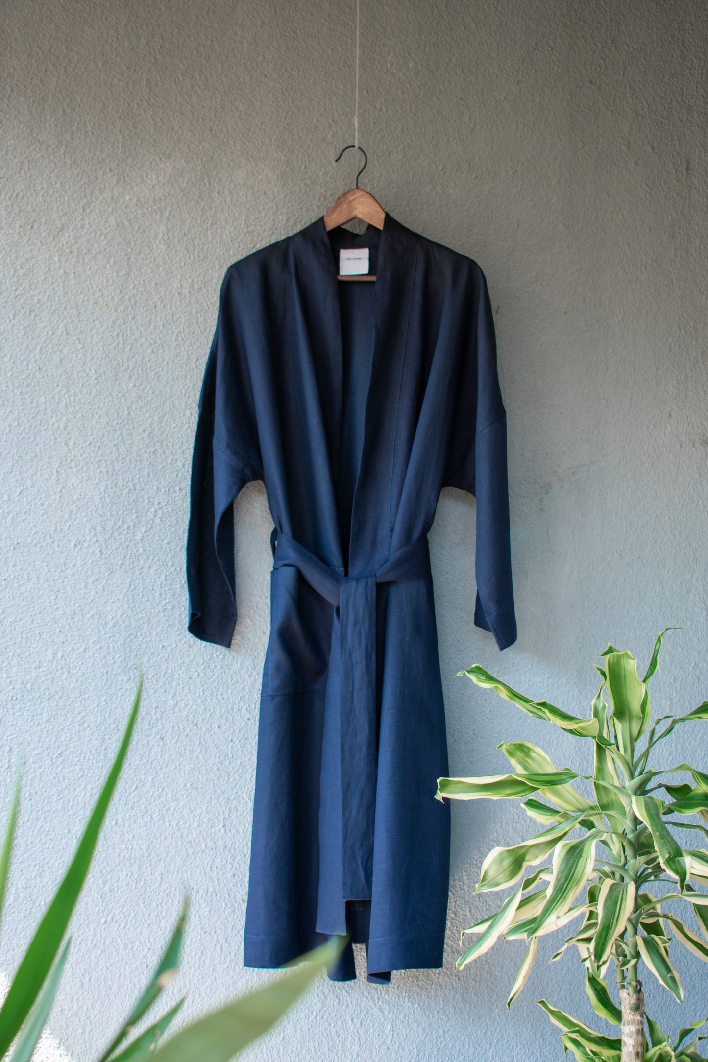 The long-sleeved, loosely tied kimono coat hangs elegantly on a hanger on a sunny grey wall. Its light midnight blue linen makes it very elegant. Fresh plants shine in the foreground.