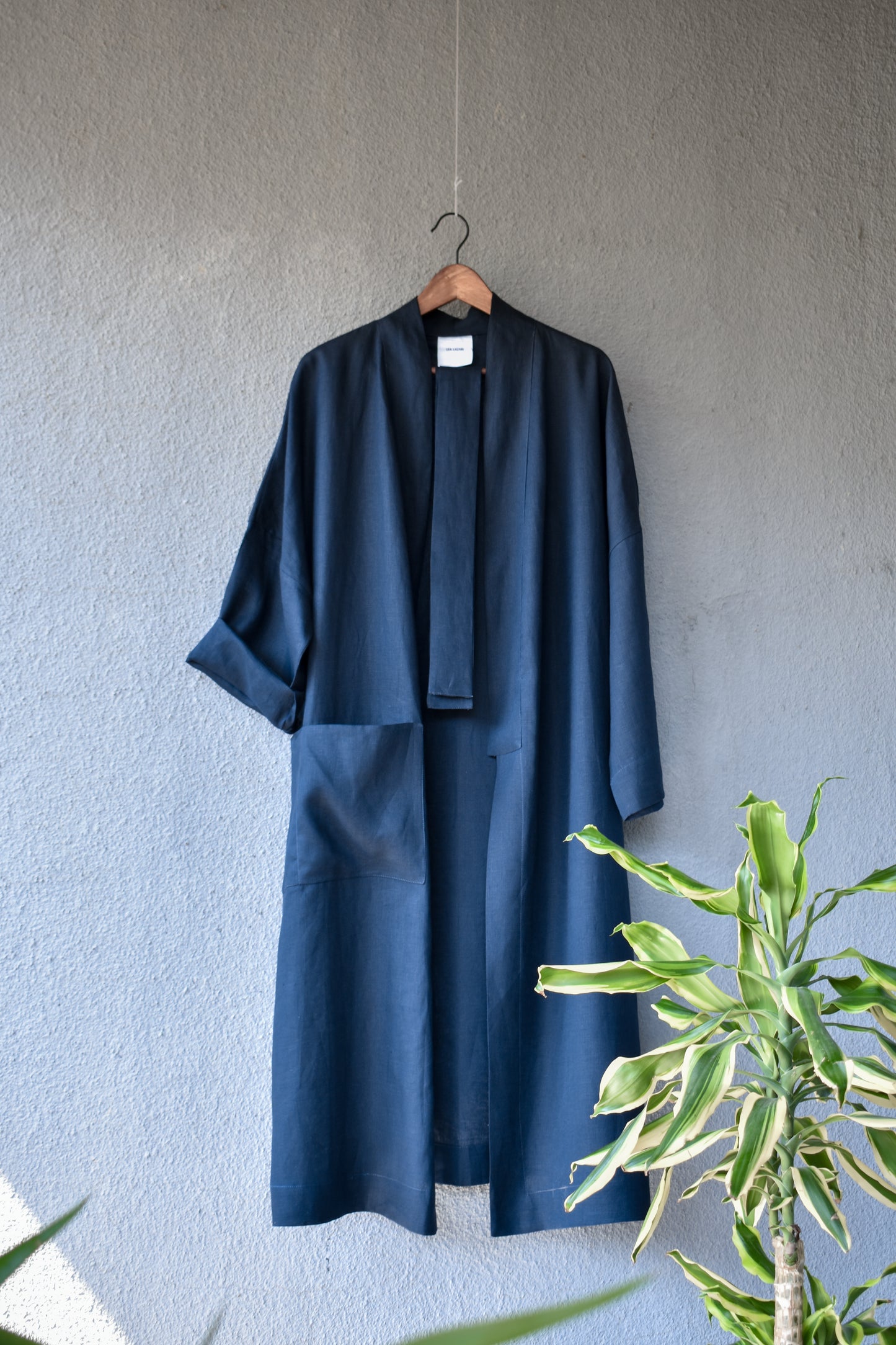 The loose dark kimono hangs open on a hanger against a light-coloured wall. The symmetrical shape is emphasised by a large side pocket, a long shawl collar and a wide belt. The linen fabric has a slight drape.