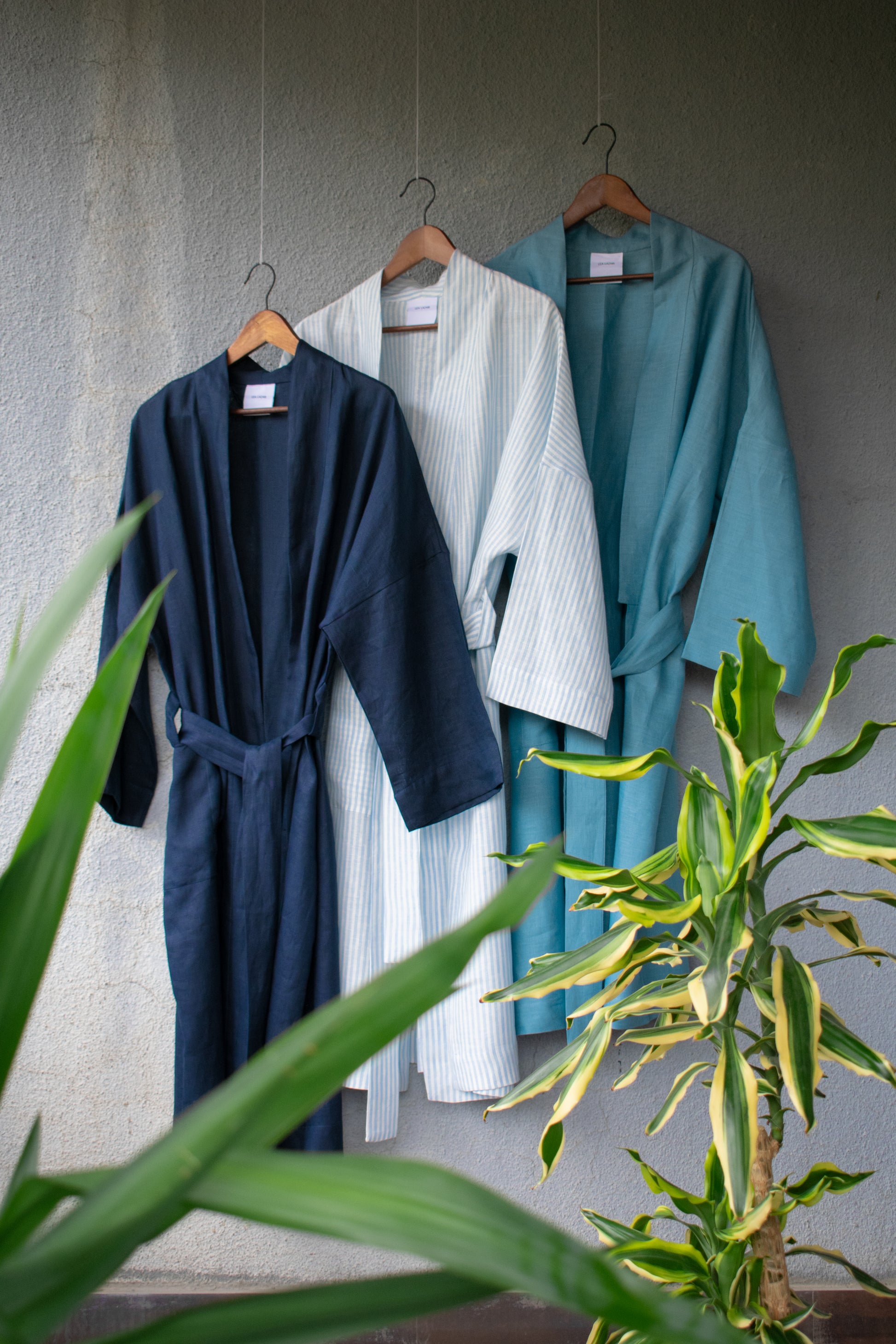 Three summery linen kimono coats in different shades of blue hang offset on a sunny wall. They are loosely tied and swing in the wind. Small green palm trees can be seen in the foreground. 